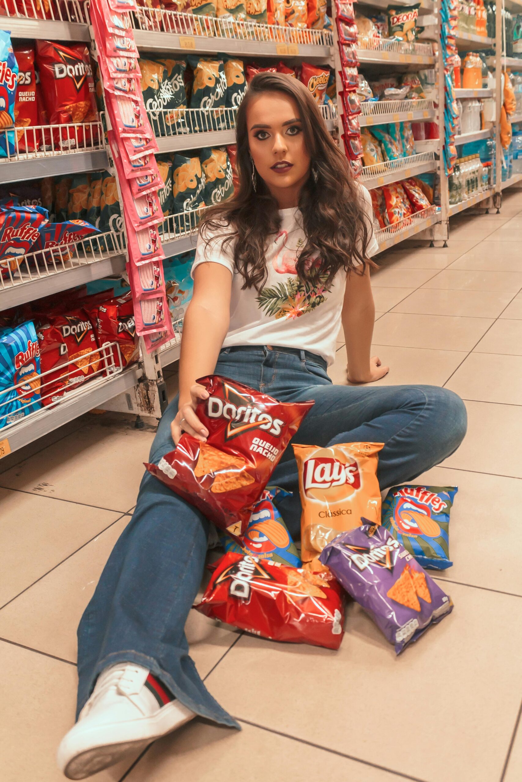 Young woman with trendy style sits on supermarket floor surrounded by snack bags.