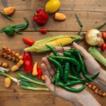 Hands holding fresh green chili peppers with assorted vegetables on wooden surface.